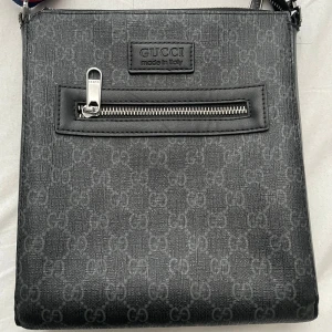 Hej jag säljer denna väskan för att jag har tänk och köpa en annan väska nu och jag har inte haft denna så länge och jag har bara rest med denna väskan. - Snygg svart axelväska från Gucci med diskret GG-mönster. Väskan har en praktisk dragkedja framtill och en justerbar axelrem i blått och rött. Perfekt för att bära dina dagliga nödvändigheter med stil.