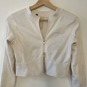 Vit ribbad jacka från Bo+tee - Snygg vit ribbad jacka från Bo+tee med dragkedja framtill. Jackan har en croppad passform och långa ärmar, perfekt för en stilren look. Den är tillverkad i ett mjukt material som ger en bekväm känsla.
