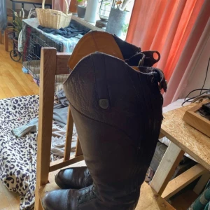 Bruna ridstövlar med dragkedja - Säljer ett par bruna ridstövlar i skinn med dragkedja baktill. Stövlarna har en klassisk design med rundad tå och dekorativa knappar upptill. Perfekta för ridning eller en stilren look. Aurora 