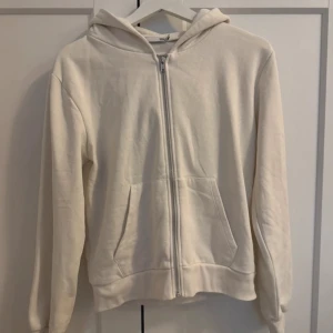 Vit hoodie med dragkedja - Säljer en stilren vit hoodie med dragkedja och huva. Tröjan har långa ärmar och praktiska fickor framtill. Perfekt för en avslappnad look. Passar till det mesta och är superbekväm!