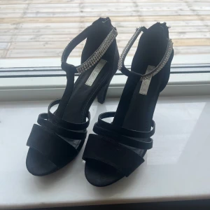 Svarta klackskor med pärldetaljer - Eleganta svarta klackskor med öppen tå och pärldetaljer på remmarna. Skorna har en dragkedja baktill för enkel på- och avtagning. Perfekta för en festlig kväll/ bal