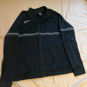 Svart träningsjacka från Nike - Säljer en svart träningsjacka från Nike med Dri-FIT-teknologi. Jackan har en dragkedja framtill och vita detaljer längs ärmarna och bröstet. Perfekt för sportiga aktiviteter eller som en stilren vardagsjacka. 🖤