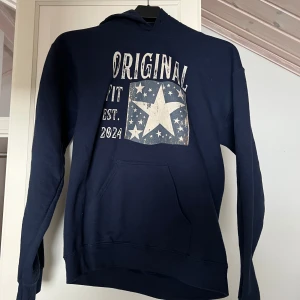 Mörkblå hoodie med stjärntryck - Säljer en mörkblå hoodie med ett stort stjärntryck och texten 'Original Fit Est. 2024' på framsidan. Tröjan har en klassisk känguruficka och en bekväm huva. Perfekt för en avslappnad stil. Nypris är på 900. Hör av er för frågor💪🏼