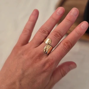 Guldfärgad ring med två ovala detaljer - Elegant guldfärgad ring med två ovala, glänsande detaljer. Denna stilrena design passar perfekt för att ge en touch av lyx till din look. Ringen har en öppen design som gör den justerbar för olika storlekar.