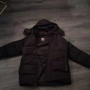 Svart pufferjacka från Canada Goose - Säljer en svart pufferjacka från Canada Goose med huva och långärmad design. Jackan har en dragkedja framtill och är perfekt för kalla dagar. Den ikoniska loggan syns på ärmen. Passar bra för vinterns alla äventyr.