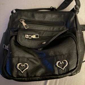 Svart axelväska med hjärtdetaljer - Snygg svart axelväska i syntetmaterial med flera dragkedjefickor och unika hjärtdetaljer i silver. Perfekt för att hålla ordning på dina saker med stil. Väskan har en justerbar axelrem för bekväm användning.