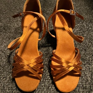 Bruna dansskor med klack - Snygga bruna dansskor med klack och korsade remmar. Skorna har en elegant design med spänne vid ankeln och är perfekta för dansgolvet. Storlek 38.