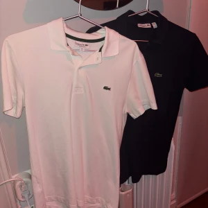 Pikétröjor från Lacoste i vitt och mörkblå - Säljer två stilrena pikétröjor från Lacoste i färgerna vitt och mörkblå. Tröjorna har klassisk krage och korta ärmar med den ikoniska krokodilloggan på bröstet. Perfekta för en avslappnad men ändå snygg look.