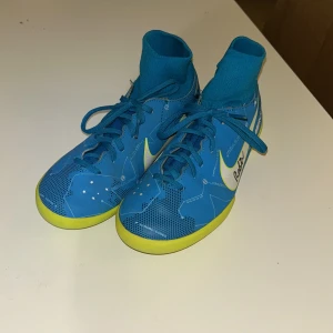 Signerade Blå Nike Mercurial fotbollsskor - Säljer ett par blå Nike Mercurial fotbollsskor med gula detaljer och snörning. Skorna har en hög ankelkrage och vit Nike-logga på sidan. Perfekta för inomhusfotboll och designade med Neymar-inspirerade detaljer. De är signerade av free skillers!