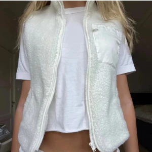 Vit fleeceväst med dragkedja - Säljer en mysig vit fleeceväst med dragkedja framtill. Västen har en praktisk bröstficka och är perfekt för kyligare dagar. Enkel och stilren design som passar till många olika outfits.