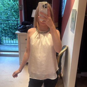 Vit siden halterneck med pärldetalj - Supersnygg vit halterneck i siden med glansig finish och pärlbroderad halsringning. Toppen är ärmlös och har en lös passform som ger en luftig känsla. Perfekt för dig som vill sticka ut med en elegant touch.