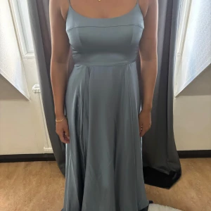 Ljusblå långklänning med smala axelband - Elegant ljusblå långklänning med smala axelband och figurnära överdel. Klänningen har en mjukt utställd kjol i glansigt material som ger ett lyxigt intryck. Perfekt för bal eller student. Enkel och stilren design utan mönster eller extra detaljer.