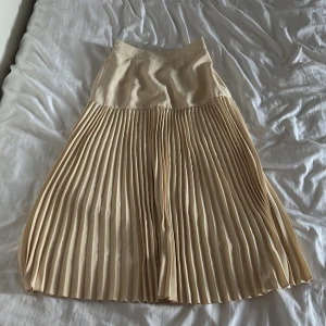 Beige plisserad långkjol - Snygg beige långkjol med plisserad nederdel och slät midja. Kjolen har en mjuk, flowy känsla och resår baktill för extra komfort. Perfekt för dig som gillar en stilren och trendig look. Passar till sneakers eller boots för en cool vibe.