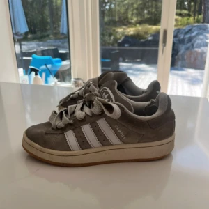 Adidas Campus grå sneakers mocka - Grå Adidas Campus sneakers i mocka med klassiska vita ränder på sidorna och vit logga på tungan och hälen. Skorna har rund tå, platt sula och breda vita snören. Perfekt för dig som gillar retrostil och streetwear.