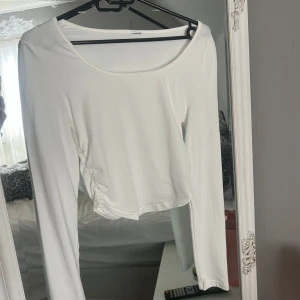 Vit croppad långärmad topp - Säljer en vit långärmad topp med croppad passform och snygg rynkdetalj på sidan. Toppen har rund halsringning och är gjord i mjukt bomullsmaterial, perfekt att styla med jeans eller kjol för en clean look.