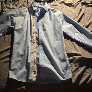 Ljusblå skjorta från Long Island - Snygg ljusblå skjorta från Long Island i slim fit-modell. Skjortan har långa ärmar och coola detaljer med mönstrad kant vid knappraden. Perfekt för dig som vill sticka ut lite extra med en klassisk men unik look.