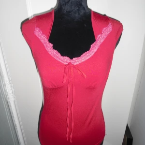 Röd topp med spets och knyt - Snygg vinröd topp med rosa spetskant vid v-ringningen och dekorativ knytning framtill. Modellen är ärmlös och har en figurnära passform som framhäver siluetten. Mörkare röd i verkligheten. Aldrig använd.