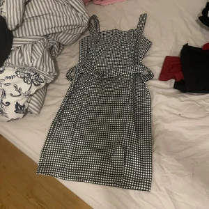 Svartvit rutig kortklänning med bälte - Supersöt svartvit rutig klänning med smala axelband och matchande bälte i midjan. Klänningen har en figurnära passform och är perfekt för dig som gillar retrovibbar och vill sticka ut lite extra.