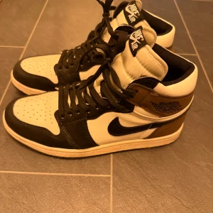 Nike Air Jordan 1 High svart/vit/brun - Säljer ett par Nike Air Jordan 1 High med svart, vit och brun färgkombination. Skorna har klassisk snörning, svart swoosh och Air Jordan-logga på sidan. Tillverkade i skinn med perforerad tåbox och högt skaft för ikonisk streetstil.