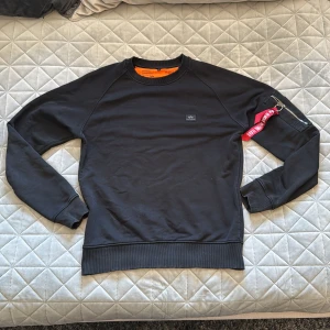 Svart Alpha Industries sweatshirt - Svart sweatshirt från Alpha Industries med klassisk rund hals och orange insida. På vänster ärm finns en dragkedja och den ikoniska röda 'Remove Before Flight'-taggen. Tröjan har ribbade muddar och en liten logotyp på bröstet. Perfekt för en stilren streetlook.