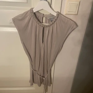 Beige snygg festtopp/blus med pärldetaljer - Supersnygg beige topp/blus med pärlbroderad halsringning och knytband i midjan. Toppen har ärmlös design och en liten öppning framtill vid halsen för extra detalj. Perfekt för dig som gillar stilrena och eleganta plagg. Pris kan diskuteras.