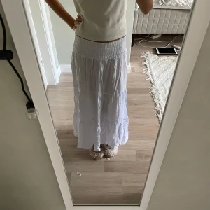 Vit långkjol från 157 - Säljer en flowy vit långkjol från 157 med smockad midja och flera volanger. Kjolen är lätt och luftig, perfekt för varma dagar. Enkel att matcha med både crop tops och t-shirts för en chill look.