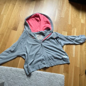 Grå hoodie från Hollister med rosa huva - Säljer en grå hoodie från Hollister i storlek L. Tröjan har dragkedja, snörning i huvan och en insida i rosa tyg. Detaljerad med ett färgglatt band längs insidan av huvan och en broderad Hollister-logga vid nederkanten. Perfekt för chill dagar.