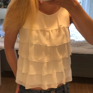 Vit volangtopp  - Supersöt vit ärmlös topp med flera lager volanger framtill. Inga defekter förutom en liten fläck längst ner under volangerna (sista bilden), men som inte märks när man har den på sig 🥰