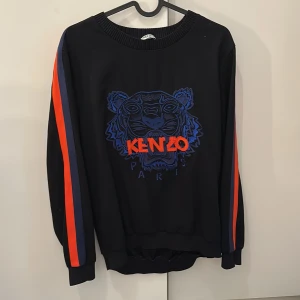 Svart Kenzo sweatshirt med tiger - Kenzo sweatshirt i svart med broderad blå tiger och röd logga på bröstet. Tröjan har långa ärmar med röda och blå ränder längs sidorna. Ribbad hals, ärmslut och nederkant. Perfekt statement-plagg för dig som gillar streetstyle.