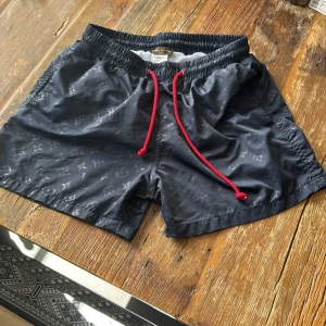 Louis Vuitton badshorts med monogram - Svarta Louis Vuitton badshorts med diskret monogrammönster och röda snören i midjan. Klassisk modell med elastisk midja, bakficka med knapp och meshfoder. Perfekta för stranden eller poolen, riktigt snygga och trendiga.