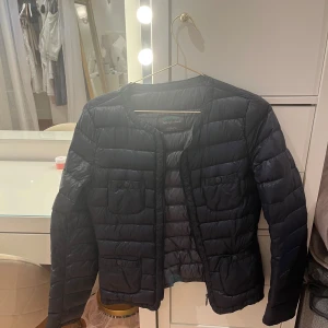 Mörkblå dunjacka från Benetton - Snygg mörkblå dunjacka från Benetton med rund halsringning och fyra fickor framtill med knappdetaljer. Jackan är quiltad och har dragkedja framtill. Perfekt för kyliga dagar och enkel att styla till olika outfits.