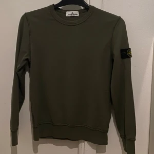 Olivgrön Stone Island sweatshirt - Snygg olivgrön sweatshirt från Stone Island Junior med klassisk rund hals och ikonisk patch. Tröjan har långa ärmar och ribbade muddar för en clean och stilren look. Storlek 142 cm men passar upp till 152 cm. Köpt för 1749 kr på Nordiska Kompaniet. Endast hämtning som gäller. Hör av er vid frågor gällande tröjan.