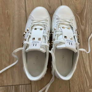 Valentino Rockstud vita sneakers - Snygga vita sneakers från Valentino med ikoniska guldiga nitar längs sidorna. Skorna har snörning, platt sula och är tillverkade i skinn för en lyxig känsla. Perfekta för dig som vill sticka ut med exklusiva detaljer.