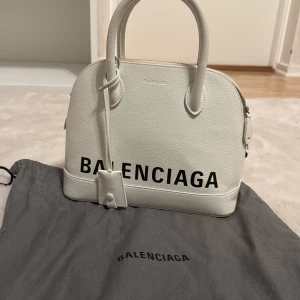 Vit handväska från Balenciaga - Säljer en snygg vit handväska från Balenciaga i skinn med svart logga framtill. Väskan har rundad form, dubbla handtag, avtagbar axelrem och dragkedja upptill. Kommer med original dustbag. Perfekt för dig som vill sticka ut med en stilren designerbag.