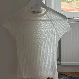 Vit stickad topp från Zara - Superfin vit stickad topp från Zara med korta ärmar och rund hals. Toppen har ett snyggt hålmönster framtill och ribbade detaljer som ger en fräsch och trendig look. Perfekt till jeans eller kjol för en chill och stilren outfit.