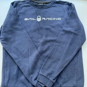 Mörkblå Sail Racing sweatshirt - Mörkblå sweatshirt från Sail Racing med vit logga framtill. Tröjan har rund hals, långa ärmar och ribbade muddar. Tillverkad i mjuk bomulls- och polyestermix, perfekt för en chill och sportig stil.