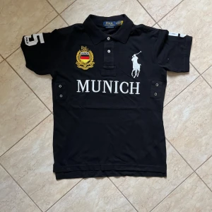 Svart pikétröja Polo Ralph Lauren Munich - Svart pikétröja från Polo Ralph Lauren med vit broderad logga, stor text 'MUNICH' och guldfärgad badge på bröstet. Nummer 5 i vitt på ryggen och ärmen. Klassisk krage och korta ärmar. Tillverkad i bomull.