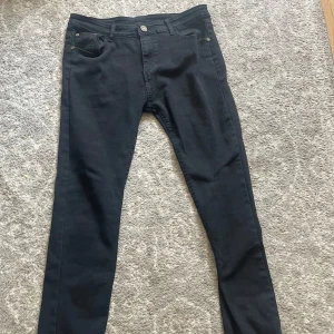 Svarta skinny jeans från Lee - Säljer ett par svarta skinny jeans från Lee med klassisk femficksdesign och snygga Lee-detaljer på bakfickan och knappen. Jeansen är i stretchigt bomullsmaterial och har en smal passform som sitter tajt längs benen. Perfekta till sneakers eller boots.