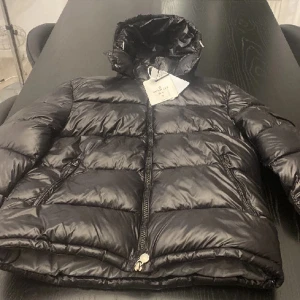  Moncler Jacka Helt ny! - Snygg svart pufferjacka från Moncler med glansig finish och huva. Jackan har två dragkedjeförsedda fickor framtill, tydlig Moncler-logga på ärmen och quiltad design. Perfekt för kalla dagar när du vill hålla stilen och värmen.