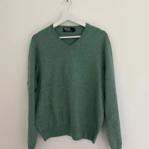 Grön stickad tröja Polo Ralph Lauren - Snygg grön stickad tröja från Polo Ralph Lauren med lila broderad logga på bröstet. V-ringad modell med långa ärmar och ribbade muddar. Storlek XL men uppskattas som M/L. 
