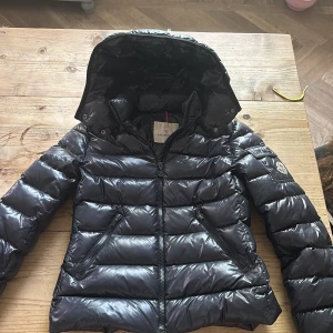 Mörkblå Moncler jacka med huva - Snygg mörkblå pufferjacka från Moncler med glansig finish och avtagbar huva. Jackan har två dragkedjefickor fram, lång ärm och klassisk Moncler-logga på ärmen. OBS trasig dragkedja längst upp som visas på sista bilden, men har själv inte ens märkt det förens man drar upp dragkedjan över de vilket jag inte brukar göra. Annars väldigt bra skick, inga andra defekter eller fläckar❤️ Passar en XS och S