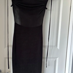 Svart långklänning med snörning - Säljer en svart långklänning med smickrande snörningsdetaljer på sidorna upptill. Klänningen har en tight passform och är tillverkad i ett glansigt, stretchigt material som ger en snygg siluett. Perfekt för dig som vill ha en stilren och elegant look. Med öppen rygg. 
