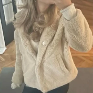 Beige teddyjacka med huva - Supermjuk och fluffig beige teddyjacka med huva och fickor framtill. Jackan har en avslappnad passform och stängs med knapp. Perfekt för dig som vill ha en cozy vibe och gillar neutrala färger. FÅR BJUDA
