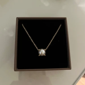 Guldhalsband med vit pärla - Elegant halsband med tunn guldfärgad kedja och en stor rund vit pärla i mitten. Perfekt för dig som gillar stilrena och klassiska smycken med en modern touch.