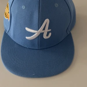 Blå keps med brodyr Los Angeles - Snygg ljusblå keps med vit broderad A på framsidan och brodyr med texten 'Los Angeles Airport 1983' i gult och rött på sidan. Justerbar snapback baktill. Kepsen är i bomull och har klassisk form med platt skärm.