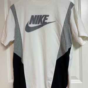Nike t-shirt med grafisk logga M - Snygg t-shirt från Nike i vitt, grått och svart med coolt grafiskt Nike-tryck på bröstet. T-shirten har korta ärmar och är gjord i mjuk bomull. Paneldesignen ger en sportig vibe och passar perfekt till streetwear eller träning.