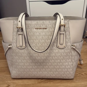 Beige Michael Kors axelväska - Snygg beige axelväska från Michael Kors med MK-logga över hela väskan. Väskan har flera fack, gulddetaljer och dragkedja upptill. Tillverkad i syntetmaterial med skinnliknande detaljer och stilren design. Perfekt för dig som vill ha en rymlig och trendig väska. Dustbag tillkommer - köpt 2022.