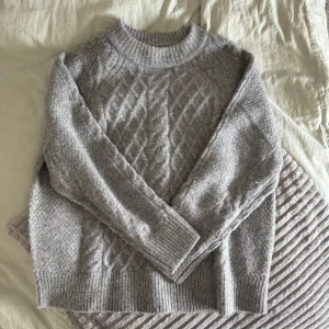 Grå stickad tröja med kabelmönster - Mysig grå stickad tröja med klassiskt kabelstickat mönster framtill. Tröjan har rund halsringning, ribbade muddar och lång ärm. Perfekt för lager-på-lager och chill dagar när du vill vara både varm och snygg.