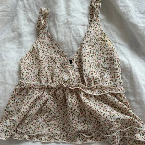 Blommigt linne med volangdetaljer - Supersöt linnetopp med småblommigt mönster i rosa, grönt och blått på ljus botten. Djup v-ringning, volangkantade axelband och fina volangdetaljer längs midjan och nederkanten. Perfekt på sommaren eller på en fika 