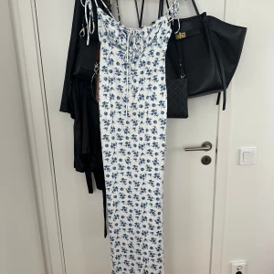 Vit långklänning med blå blommor - Supersöt långklänning i vitt med blått blommönster och smala axelband med knyt. 